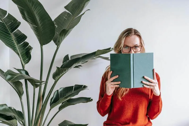 uma moça de pé segurando um livro verde com apenas os olho a vista, e uma planta ao lado para exemplificar o post de livros parecidos com verity